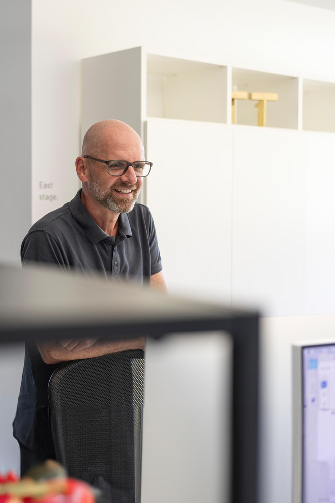 Ein lächelnder Mann mit Glatze und Brille steht an einem Schreibtisch in einem modernen Büro. Er trägt ein graues Polo-Shirt und wirkt entspannt und freundlich. Im Hintergrund sind weiße Schränke zu sehen, und an der Wand steht der Text East stage. Der Mann beobachtet aufmerksam eine Situation oder Interaktion, während er sich an einem Stuhl anlehnt.