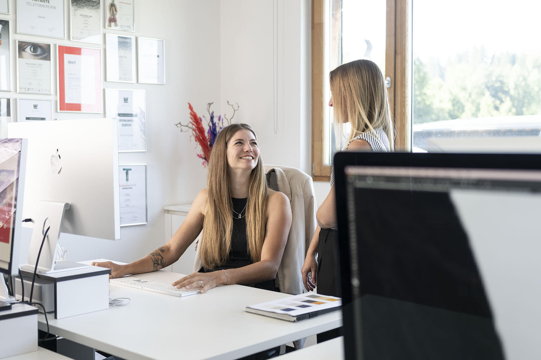 Zwei Frauen sitzen in einem modernen Büro und lächeln, während sie miteinander sprechen. Im Hintergrund sind zahlreiche Zertifikate und Auszeichnungen an der Wand angebracht. Vor der ersten Frau steht ein iMac, und der Fokus auf dem Bild zeigt die freundliche Atmosphäre des Büros