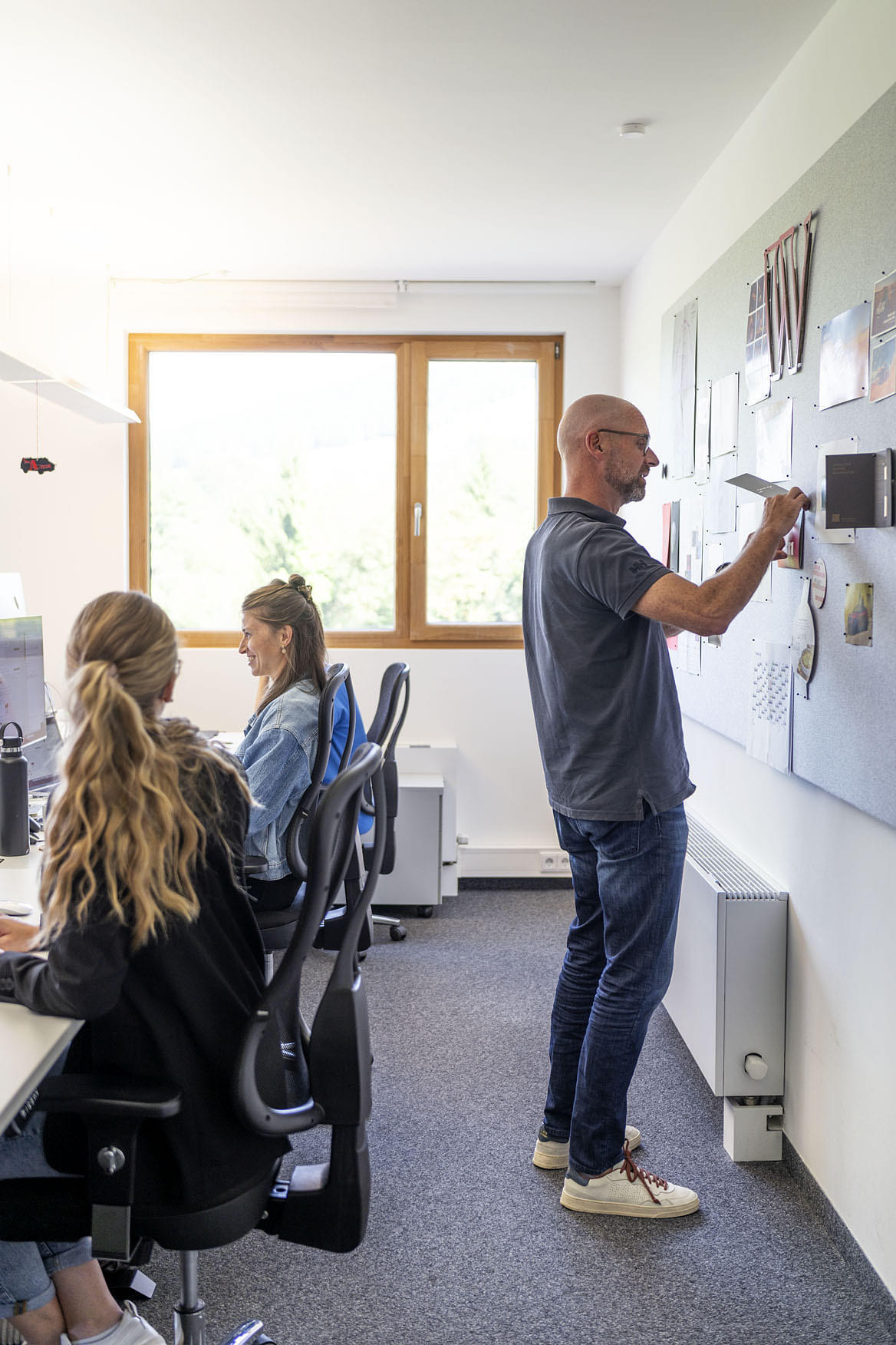 Ein kreatives Büro mit zwei Mitarbeiterinnen, die an ihren Schreibtischen sitzen und an Computern arbeiten. Im Vordergrund ist eine Frau mit langen, gewellten Haaren zu sehen, während eine andere Frau im Hintergrund aufblickt. Rechts im Bild steht ein Mann mit Glatze, der Notizen an eine Wandtafel mit verschiedenen Informationen und Bildern anheftet. Große Fenster sorgen für natürliches Licht im Raum.