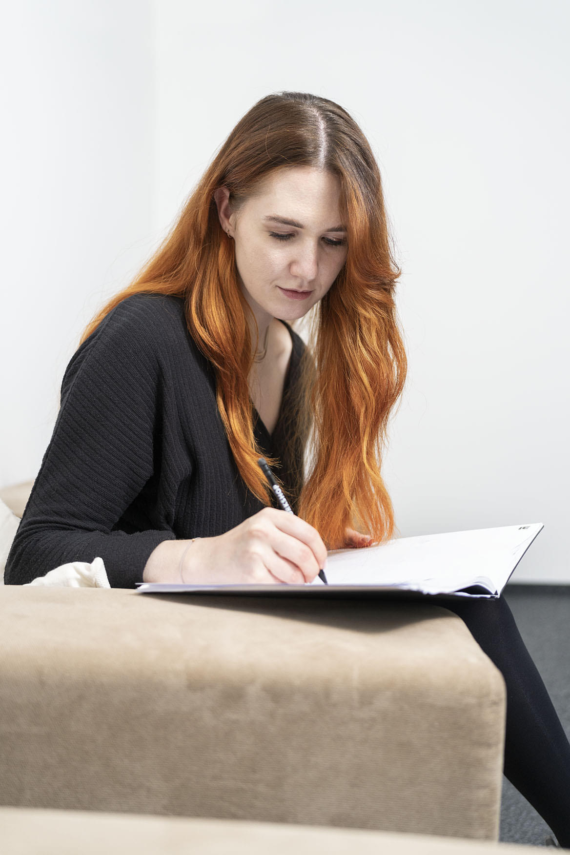 Eine Frau mit rotbraunen Haaren sitzt auf einem beigen Sofa und zeichnet konzentriert in ein Notizbuch.