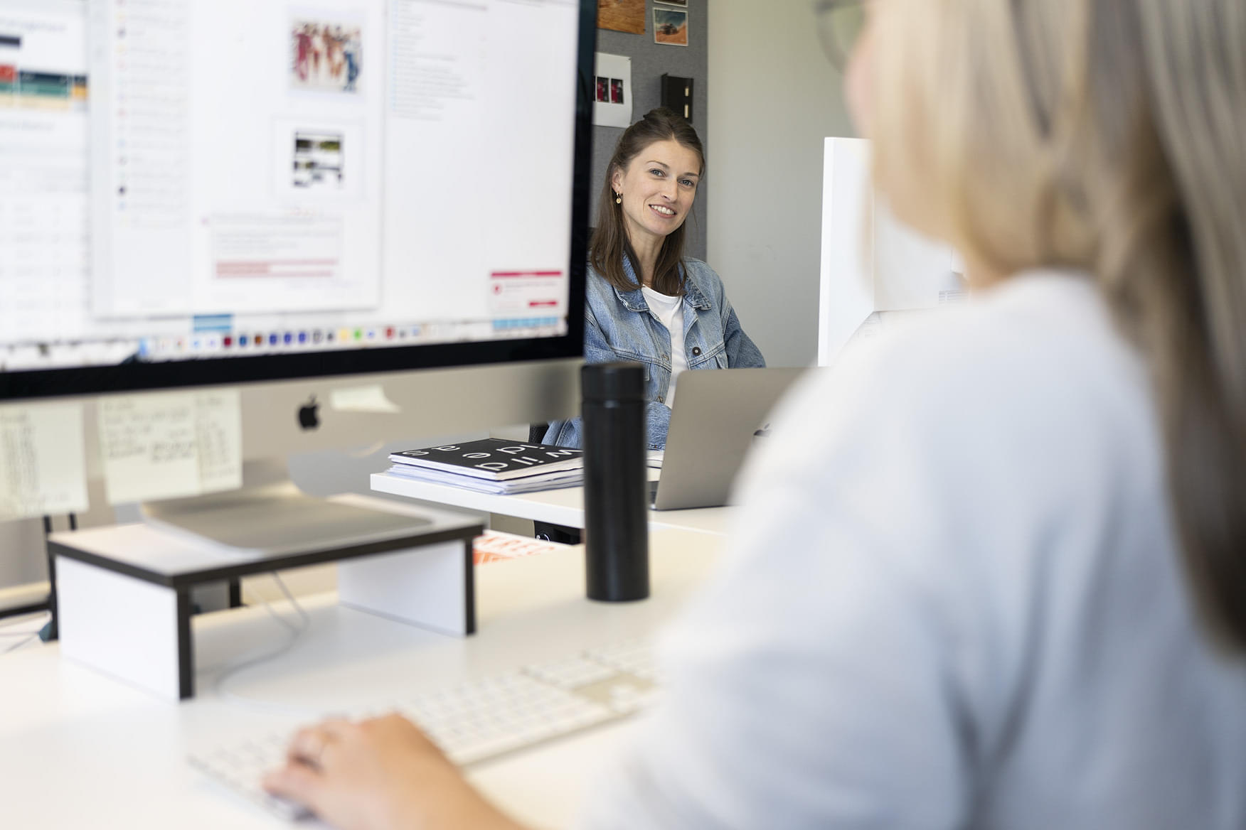 Zwei Frauen arbeiten in einem modernen Büro. Die Frau im Vordergrund mit blonden, langen Haaren und einem grauen Oberteil ist dabei, auf eine Computermaus zu klicken. Auf dem Bildschirm vor ihr sind verschiedene Programme und Anwendungen sichtbar. Im Hintergrund sitzt eine weitere Frau mit langen, dunklen Haaren in einem Jeansjacke, die lächelt und Blickkontakt mit der anderen Frau hat. 