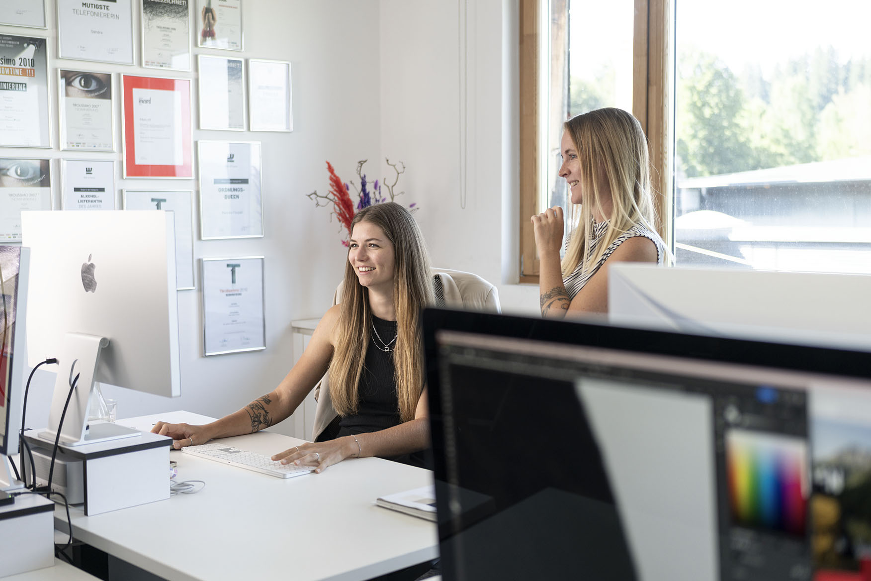 Zwei Frauen sitzen in einem modernen Büro und lächeln, während sie miteinander sprechen. Im Hintergrund sind zahlreiche Zertifikate und Auszeichnungen an der Wand angebracht. Vor der ersten Frau steht ein iMac, und der Fokus auf dem Bild zeigt die freundliche Atmosphäre des Büros.